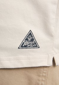 Beige katoenen T-shirt met een geribbelde textuur, voorzien van een marineblauwe driehoekige logo-patch met een geit en de tekst "Petrol" dicht bij de zoom.