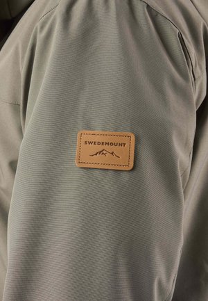 Leren patch in tan met de tekst "Swedemount" en een berglogo, vastgestikt op de bovenkant van de mouw van een olijfgroene jas.