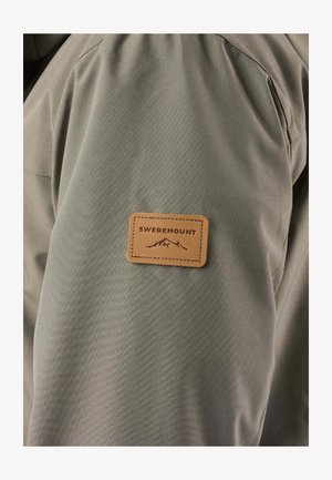 Leren patch in tan met de tekst "Swedemount" en een berglogo, vastgestikt op de bovenkant van de mouw van een olijfgroene jas.