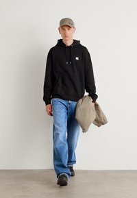 Zwarte hoodie met kangoeroezak en trekkoord, blauwe losvallende jeans, grijze pet en een beige jas in de hand, gedragen met zwarte schoenen.