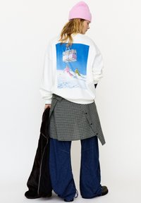 White sweatshirt featuring a colorful skiing graphic on the back, paired with a checkered skirt overlay and loose blue denim pants.