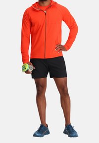 Homme en veste zippée orange vif, short noir, chaussures de course bleues, tenant une bouteille d'eau avec un bracelet jaune sur un fond uni.