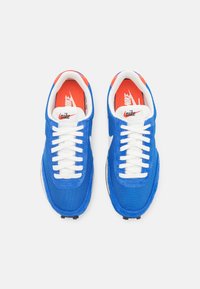 Blue mesh and suede sneakers with white laces, a red and white insole, and a signature logo on the tongue. Rounded toe and textured sole.