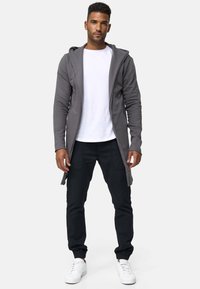 Sudadera gris ligera con frente abierto, con una camiseta blanca debajo, combinada con pantalones jogger negros y zapatillas blancas. Textura de tela suave.