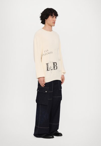 Pull à manches longues couleur écru avec inscription "LES ENFANTS L.B." ; associé à un pantalon cargo en denim foncé avec plusieurs poches et détails de couture.