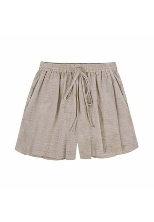 Kobieta z długimi blond włosami, ubrane w czarny crop top i beżowe szorty, poprawiająca koszulę stojąc na zewnątrz o zachodzie słońca.