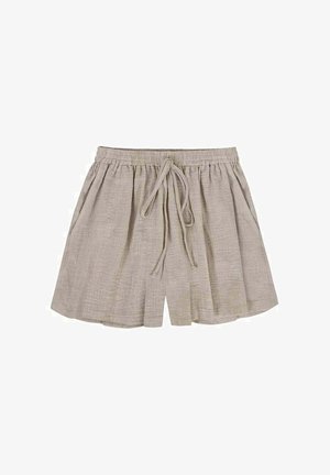 Woman with long blonde hair wearing a black crop top and beige shorts, adjusting her shirt while standing outdoors at sunset.