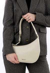 Cremfarbene Ledertasche mit geschwungener Form, strukturiertem Finish und kurzem Griff mit goldfarbener Hardware. Dezente Markenkennzeichnung auf der Vorderseite.