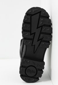 Suela de bota de goma negra con dibujo texturizado y diseño destacado de rayo. Presenta patrones en relieve y un logo de marca circular.