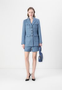 Blazer in tweed blu con quattro tasche e chiusura a bottoni, abbinato a pantaloni corti coordinati. Gli accessori includono una borsa blu e décolleté nere.