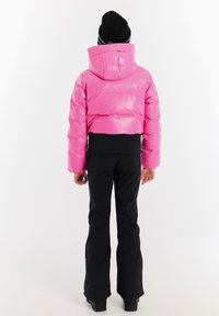 Rosa isolierte Pufferjacke mit Kapuze, mit einem verkürzten Design. Kombiniert mit schwarzen Schneehosen und schwarzen Stiefeln.