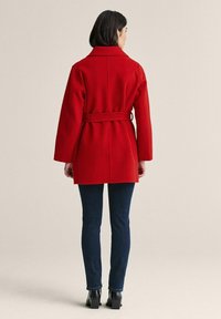 Punainen villatakki, jossa on vyötärövyö, pitkät hihat ja kauluskäänteet. Päällä tummien farkkujen ja mustien, paksupohjaisten saappaiden kanssa.