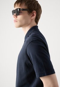 Polo azul-marinho feito de tecido suave, com mangas curtas e colarinho, combinado com óculos de sol retangulares pretos com detalhes em dourado.