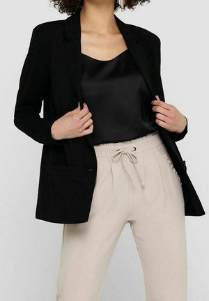 Persoon draagt zwarte blazer en zwarte satijnen top met lichtbeige broek met trekkoorden, poseert met handen die de randen van de blazer vasthouden.