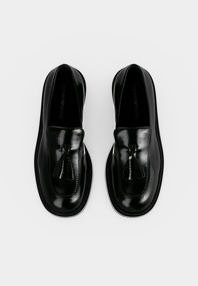Ζευγάρι μαύρα γυαλιστερά δερμάτινα loafers με φούντες, στρογγυλεμένες μύτες και χαμηλά τακούνια, εμφανιζόμενα από πάνω σε λευκό φόντο.