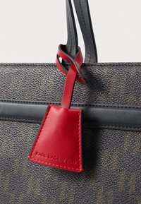 Zbliżenie na teksturowaną, ciemnobrązową torebkę z czarnymi wykończeniami oraz czerwoną, skórzaną metką z wytłoczonym napisem "KARL LAGERFELD", zawieszoną na czarnym uchwycie.