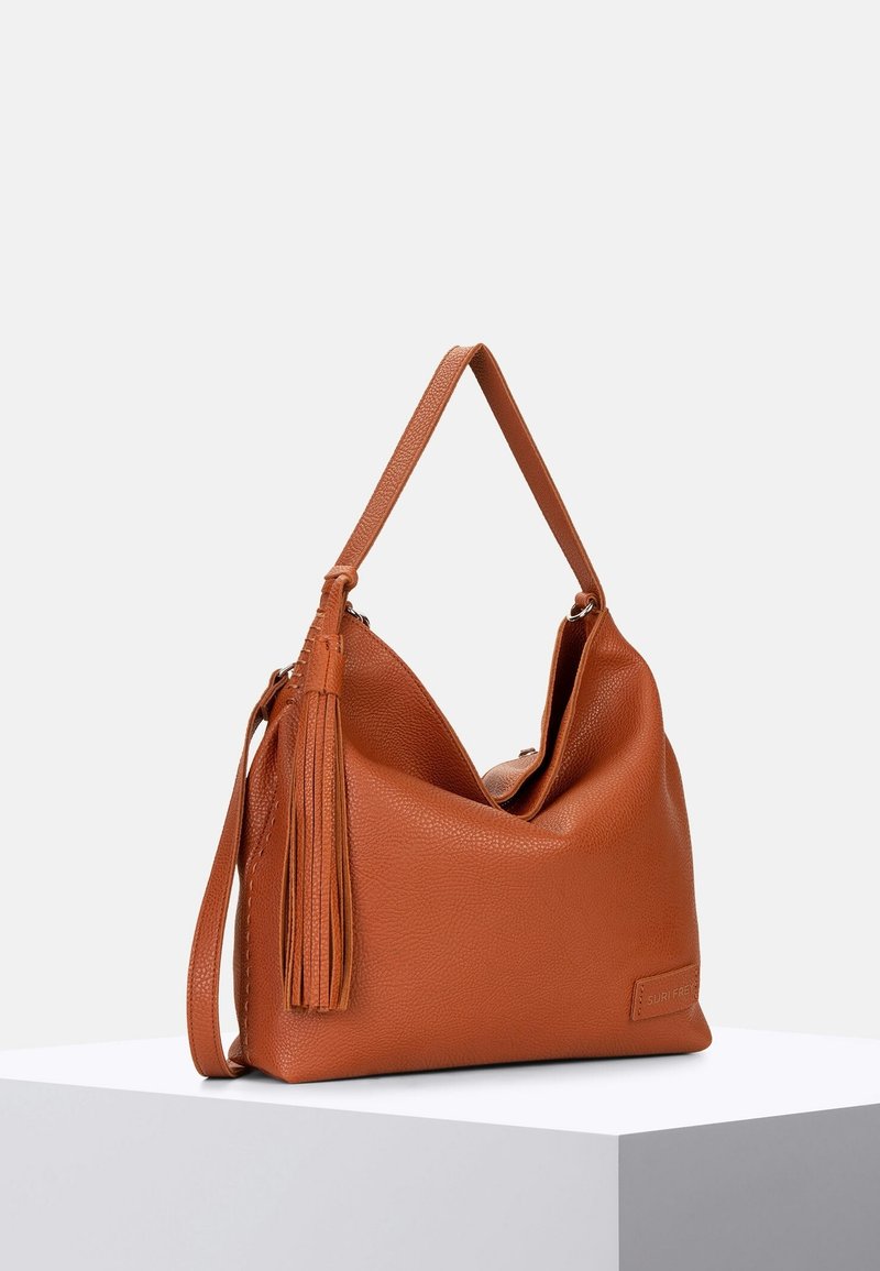 Sac à main en cuir marron texturé avec une bandoulière et un long détail de pampille, posé sur un piédestal blanc sur un fond uni.