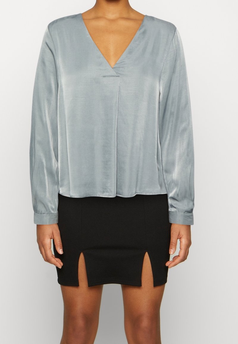Femme portant un chemisier en satin gris clair à manches longues et col en V, ainsi qu'une jupe noire avec deux fentes à l'avant.