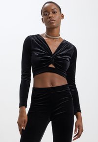 Top corto in velluto nero con maniche lunghe, scollatura a V profonda, dettaglio anteriore annodato e vita con taglio. Abbinato a pantaloni neri aderenti coordinati.