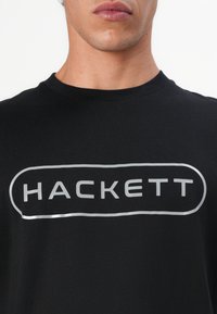 Juodas medvilnės marškinėliai su sidabriniu "HACKETT" logotipu apvalaus stačiakampio kontūre, apvalia iškirpte ir trumpomis rankovėmis.