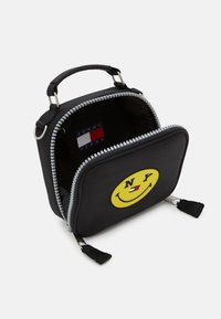 Borsa in tessuto nera con un volto sorridente giallo e il testo "NY", dotata di chiusura con zip, manico e etichetta interna con la scritta "TOMMY JEANS."