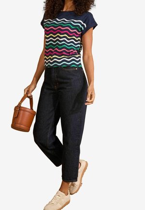 Marineblå t-shirt med flerfarvet bølget mønster, kombineret med mørk denimjeans. Holder en brun spandtaske og har beige sneakers på.