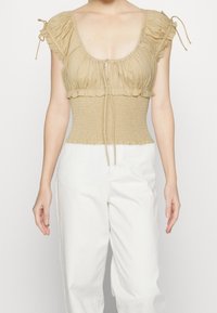 Béžový top s kulatým výstřihem, nařasenými rukávy a zbrázděnou beden. Obsahuje šňůrky pro úpravy a strukturovanou látku.