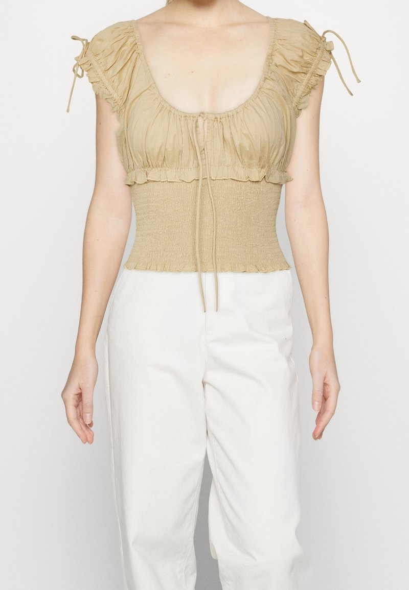 Béžový top s kulatým výstřihem, nařasenými rukávy a zbrázděnou beden. Obsahuje šňůrky pro úpravy a strukturovanou látku.