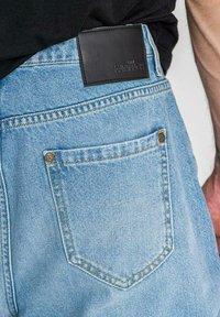 Vaaleansiniset farkut, joissa on musta nahkalaikka vyötäröllä. Niissä on yksi takatasku, jossa on keltainen ompelu ja nappi.