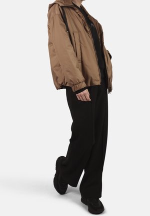 Persoon die een bruine capuchonjas met elastische manchetten, zwarte broek en zwarte schoenen draagt, staand tegen een effen witte achtergrond.