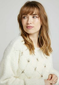 Pull en laine de couleur crème, doux et duveteux, avec un motif en losanges, orné de perles en forme d'étoile dorées et de sequins. Texture douce, coupe décontractée.