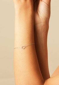 Pols met verfijnde zilveren kettingarmband met een klein, open hartvormig bedeltje tegen een beige achtergrond.