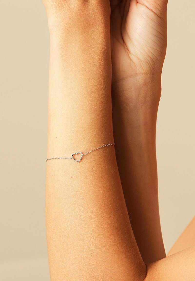 Pols met verfijnde zilveren kettingarmband met een klein, open hartvormig bedeltje tegen een beige achtergrond.