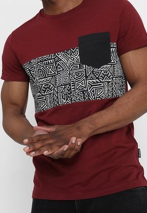 Vinrød T-shirt med en sort lomme, detaljeret med et geometrisk hvidt mønster over brystet, korte ærmer og en afslappet pasform.