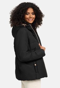 Black puffer jacket with a hood, zip front, and button accents. Features quilted design and a relaxed fit, paired with cream-colored pants.