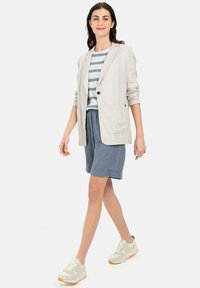 Lichtbeige linnen blazer over een blauw-witte gestreepte shirt, gecombineerd met grijze shorts en witte sneakers met lichtbruine accenten.