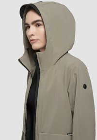 Jachetă ușoară, de culoare khaki, cu glugă, închidere cu fermoar și buzunare laterale. Prezintă un interior negru și un accent cu logo circular. Textură netedă.