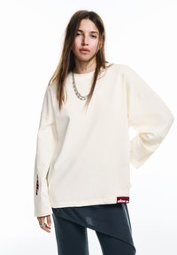 Túlméretezett krémszínű pamut pulóver lehajtott vállakkal, bordó oldalszegélycímkével és laza szabással. Sötétkék nadrággal párosítva.