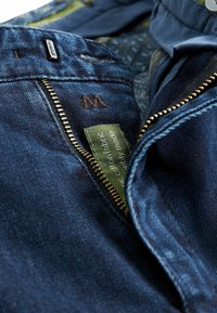Dark blue denim jeans with a gold zipper, green fabric label, and an embroidered "M." Textured surface with subtle fading details.