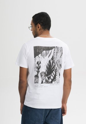 Man met een bril die een wit t-shirt draagt met een zwart-wit abstract bos- en bladdesign op de rug, gecombineerd met een blauwe spijkerbroek.
