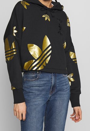 Sweat à capuche noir raccourci avec grands logos feuille métalliques dorés, associé à un jean en denim bleu, présenté des épaules aux hanches.