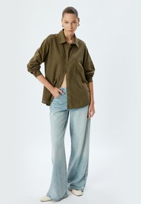 Camicia oversized verde oliva con bottoni, jeans ampi blu chiaro sbiaditi e scarpe bianche a punta, stilizzati con maniche arrotolate e dettagli discreti.