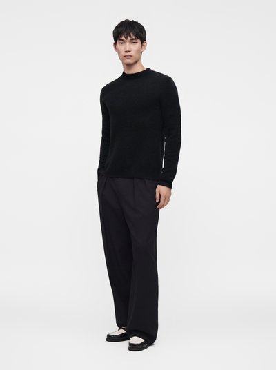 Man standing wearing a black long-sleeve sweater, loose black pants, and black and white loafers on a plain white background.