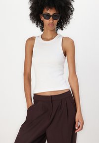 Top alb cu dungă, cu un gât rotund, asortat cu pantaloni largi, cu talie înaltă, de culoare maro închis. Modelul poartă ochelari de soare negri cu forme de ochi de pisică.