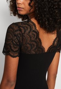 Schwarzes Kleid mit einer Spitzenpasse mit floralen Mustern und gewellten Kanten. Das Design betont eine taillierte Silhouette und strukturierte Details.