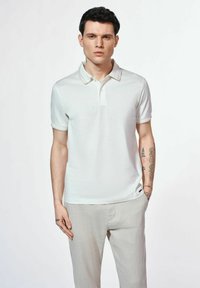 Biała koszulka polo z krótkim rękawem, z klasycznym kołnierzykiem i subtelnymi szczegółami tekstur. Nosi ją osoba w jasnobeżowych spodniach, ukazując tatuaże.