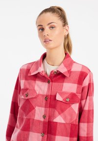 Camicia di flanella a quadri rossi e rosa con colletto, due tasche anteriori e bottoni marroni. Il tessuto ha un aspetto morbido e strutturato.