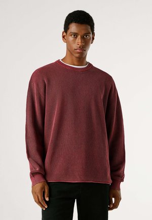 Felpa di maglia color borgogna con trama a coste, scollo rotondo e maniche lunghe. Pantaloni neri completano il look casual.