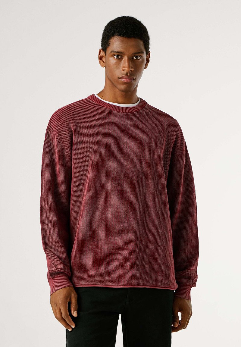 Dunkelroter Strickpullover mit geripptem Muster, Rundhalsausschnitt und langen Ärmeln. Schwarze Hose vervollständigt das legere Outfit.