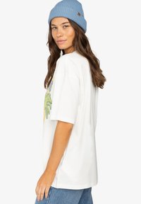 Bílý oversized tričko s barevným grafikou palmy, doplněné světle modrým žebrovaným čepicí. Model má na sobě světlé modré džíny.